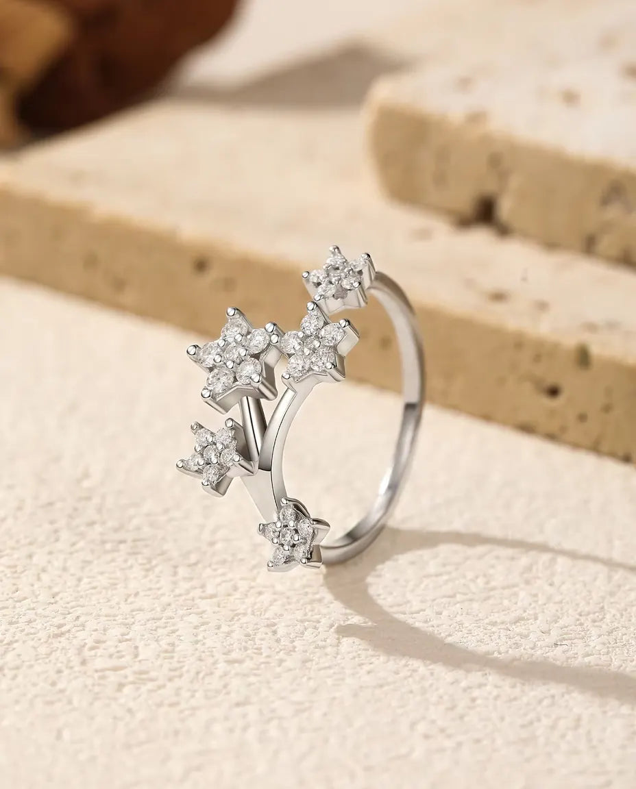 Åben ring i 925 sterling sølv med fem små stjerner af moissanite-sten på to grene, på råhvid puds ved en tørret rose.