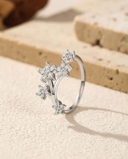Åben ring i 925 sterling sølv med fem små stjerner af moissanite-sten på to grene, på råhvid puds ved en tørret rose.