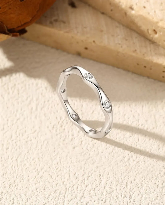 Bølget ring i 925 sterling sølv med små moissanite-sten, skråt på råhvid puds foran lyse travertintrin og tørrede blomster.