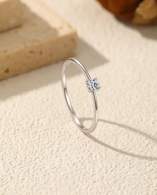 Smal ring i 925 sterling sølv med én lille moissanite i fire kløer, stillet skråt på råhvid puds foran lyse travertintrin.