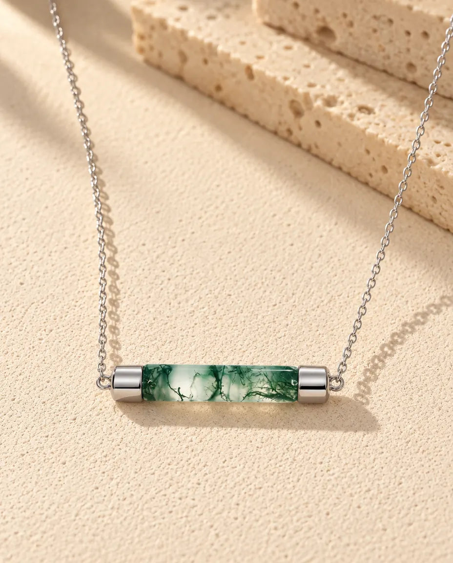 Halskæde i sølv med en grøn cylinder af moss agate og en sølvhætte i hver ende på beige puds foran travertin i varmt sidelys.