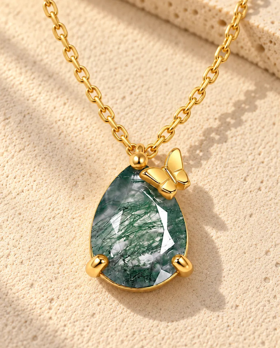 Guldbelagt halskæde med en grøn moss agate-dråbe og en lille sommerfugl øverst til højre på beige puds foran travertin.