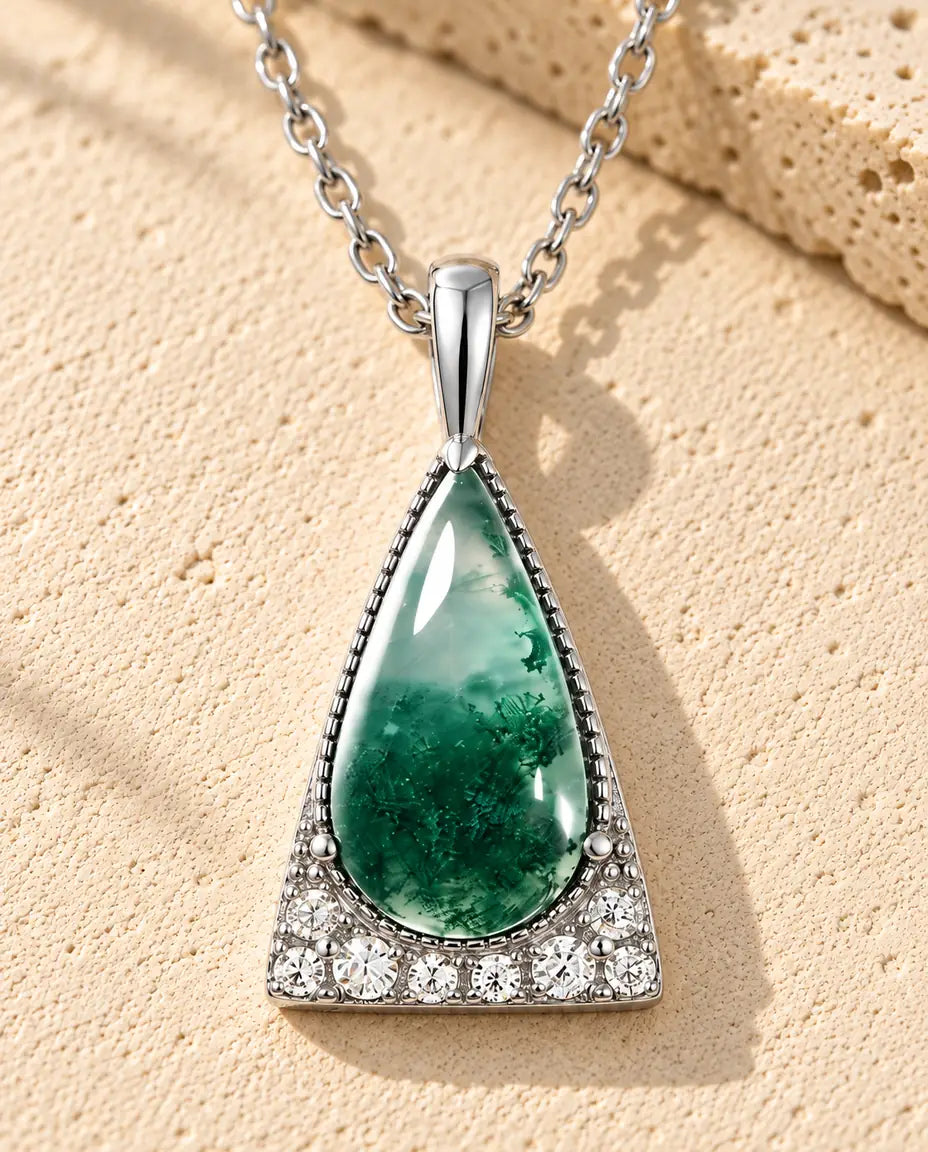 Sølvhalskæde med en grøn moss agate-dråbe i en trekantet ramme og små hvide zirkonia forneden på beige puds foran travertin.