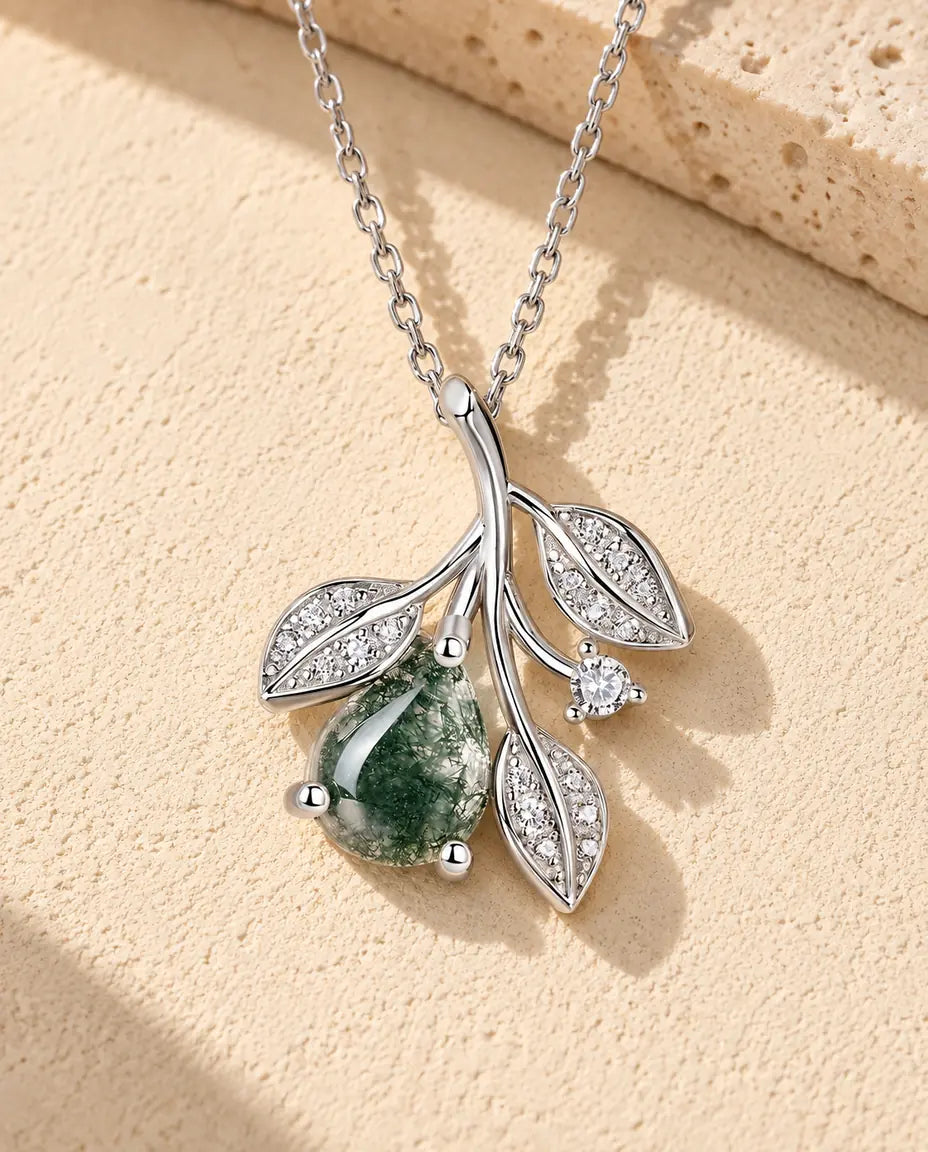 Halskæde i sølv med en gren, tre glitrende blade og en grøn dråbe af moss agate på beige puds foran travertin i sidelys.