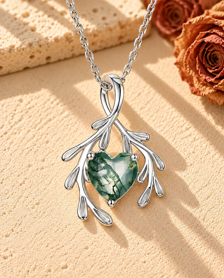 Sølvhalskæde med et facetteret moss agate-hjerte mellem to grene med små blade på beige puds foran travertin.