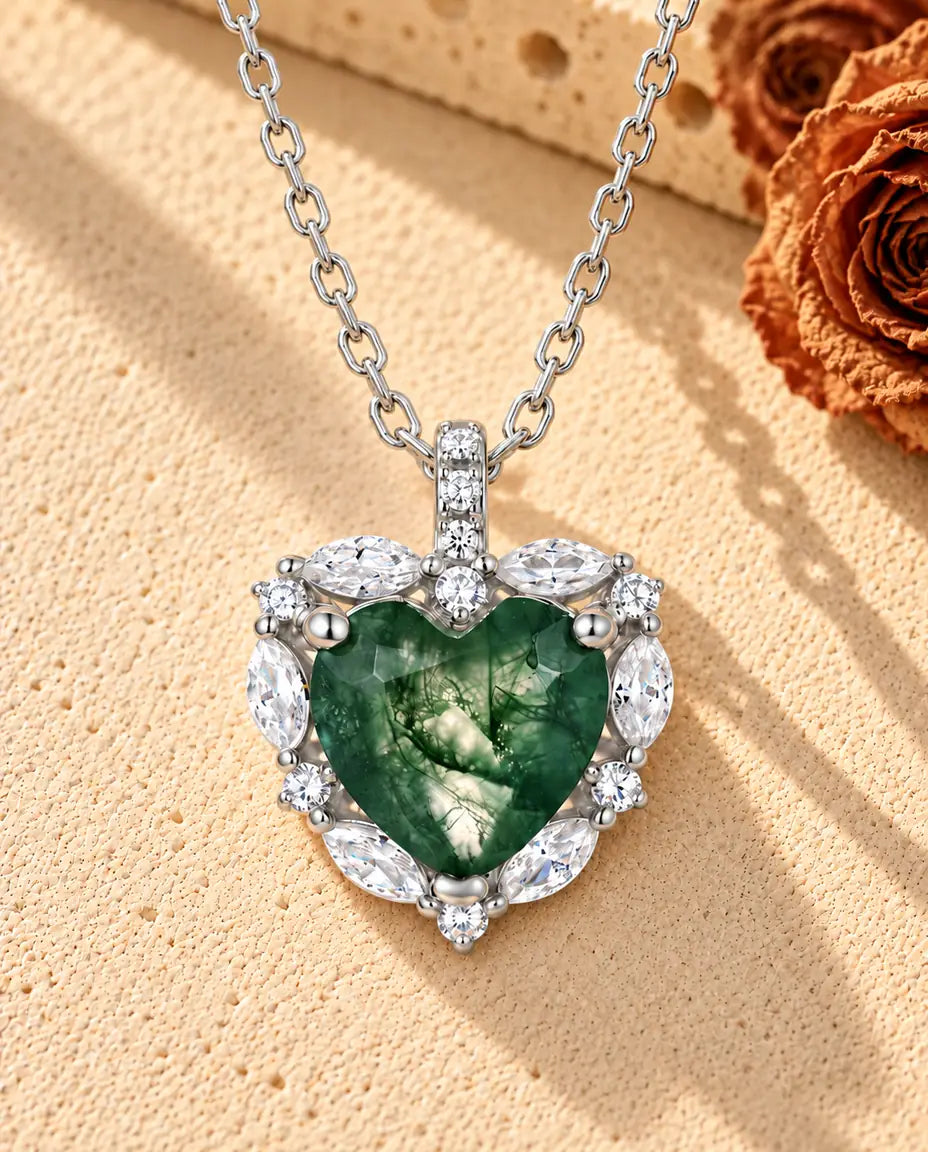 Sølvhalskæde med et moss agate-hjerte i en krans af små klare sten på beige puds foran travertin og tørrede roser.