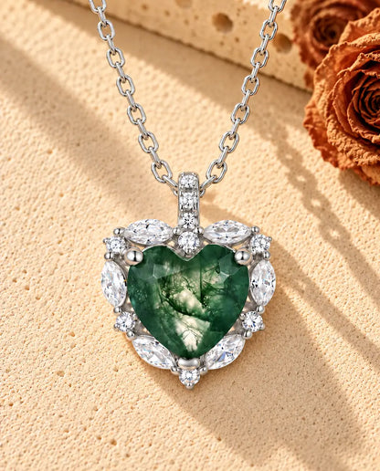 Sølvhalskæde med et moss agate-hjerte i en krans af små klare sten på beige puds foran travertin og tørrede roser.