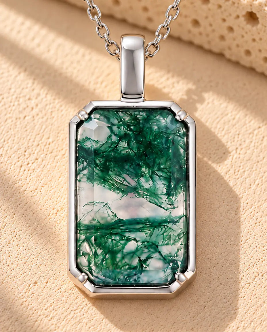 Sølvhalskæde med en klar, grøn rektangulær moss agate i en smal fatning under en bred øsken på beige puds foran travertin.