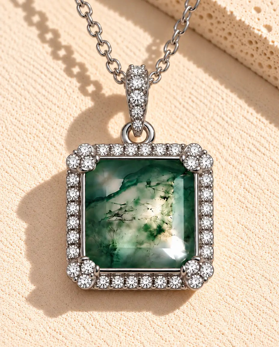 Halskæde i sølv med en grøn kvadratisk moss agate i en firkantet halo af små hvide zirkonia på beige puds foran travertin.
