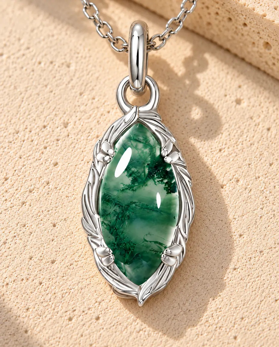 Sølvhalskæde med en grøn marquiseformet moss agate-cabochon i en ramme af sølvblade og ranker på beige puds foran travertin.