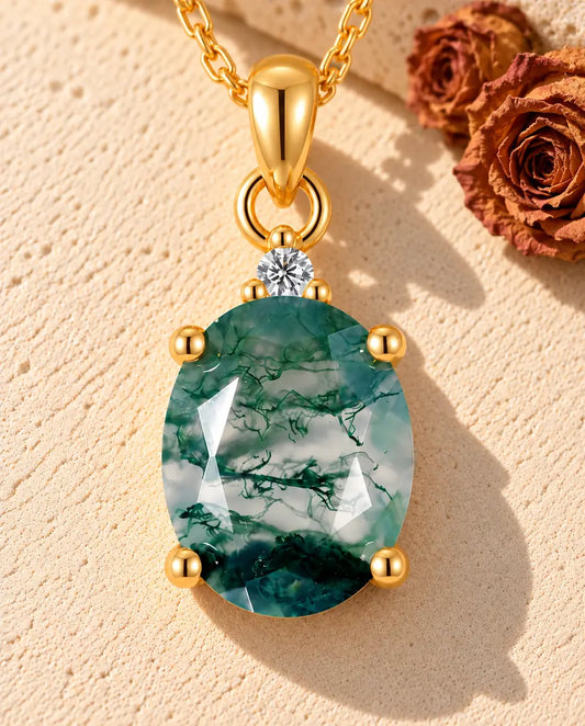 Guldbelagt halskæde med en grøn oval moss agate i fire kløer og en lille hvid zirkonia på beige puds foran travertin.
