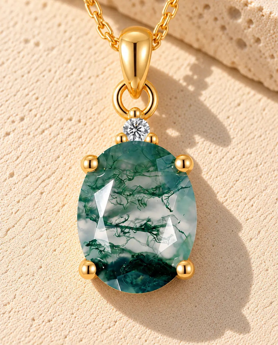 Guldbelagt halskæde med en grøn oval moss agate i fire kløer og en lille hvid zirkonia på beige puds foran travertin.