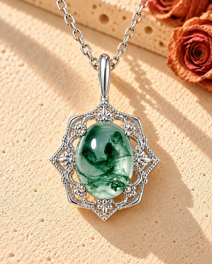 Vintage halskæde i sølv med en oval moss agate i gennembrudt blonderamme på beige puds foran travertin og en tørret rose.