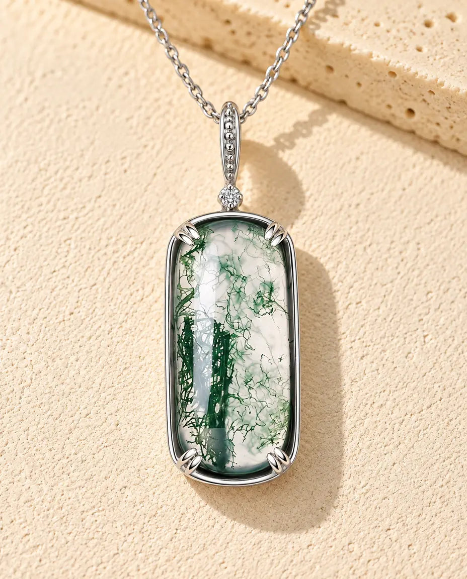Halskæde i sølv med en lang, klar rektangulær moss agate med fine grønne mostegninger på beige puds foran travertin.