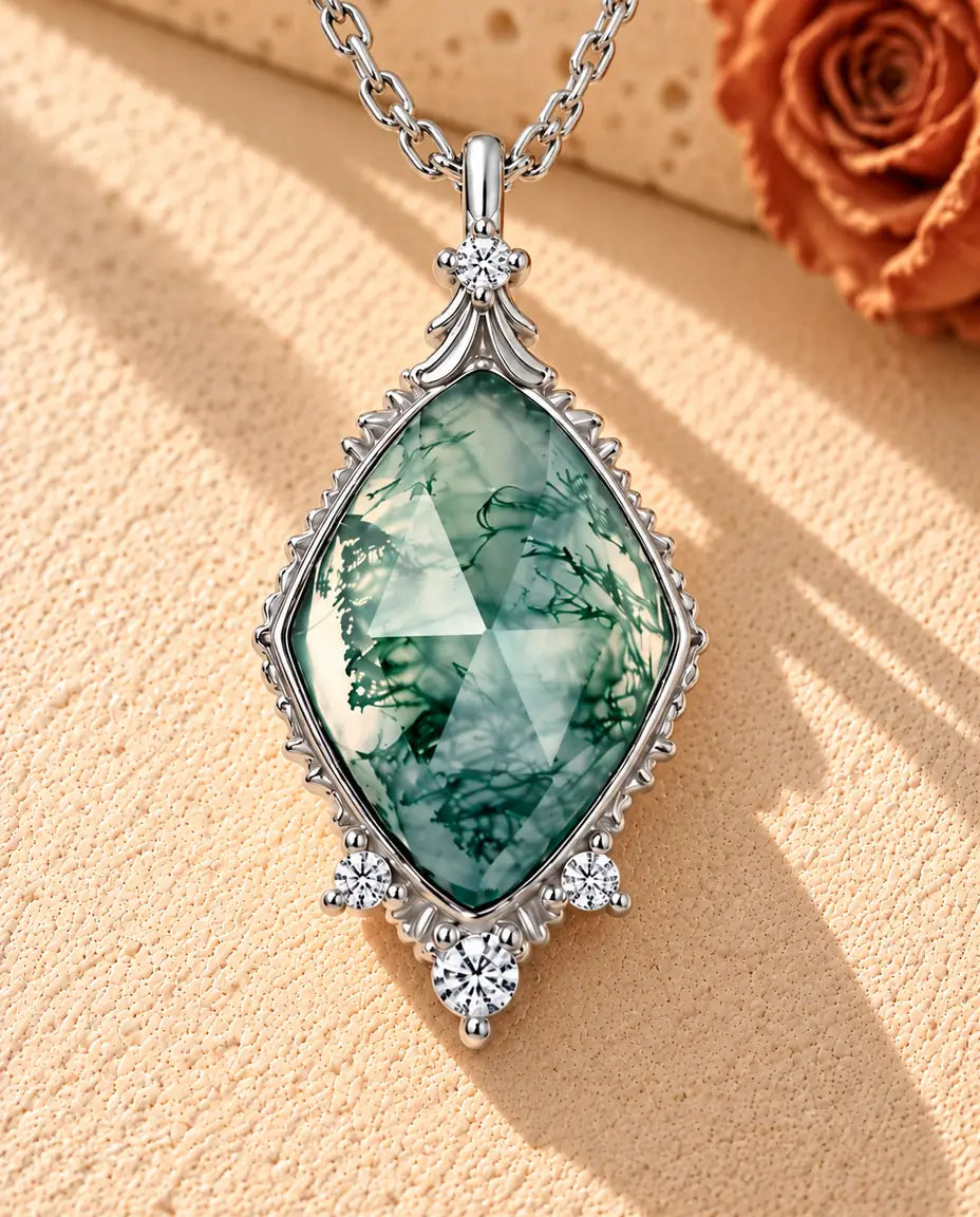 Sølvhalskæde med en rosenslebet moss agate-rombe i en ciseleret vintagefatning på beige puds foran travertin.