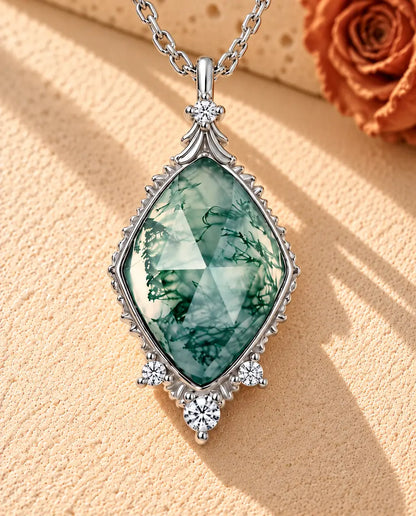 Sølvhalskæde med en rosenslebet moss agate-rombe i en ciseleret vintagefatning på beige puds foran travertin.