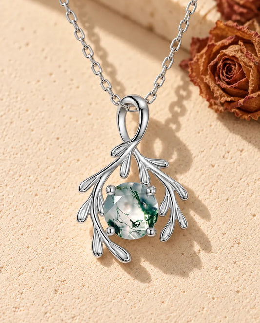 Halskæde i sølv med en grøn rund moss agate mellem to sølvgrene med små runde blade på beige puds foran travertin.
