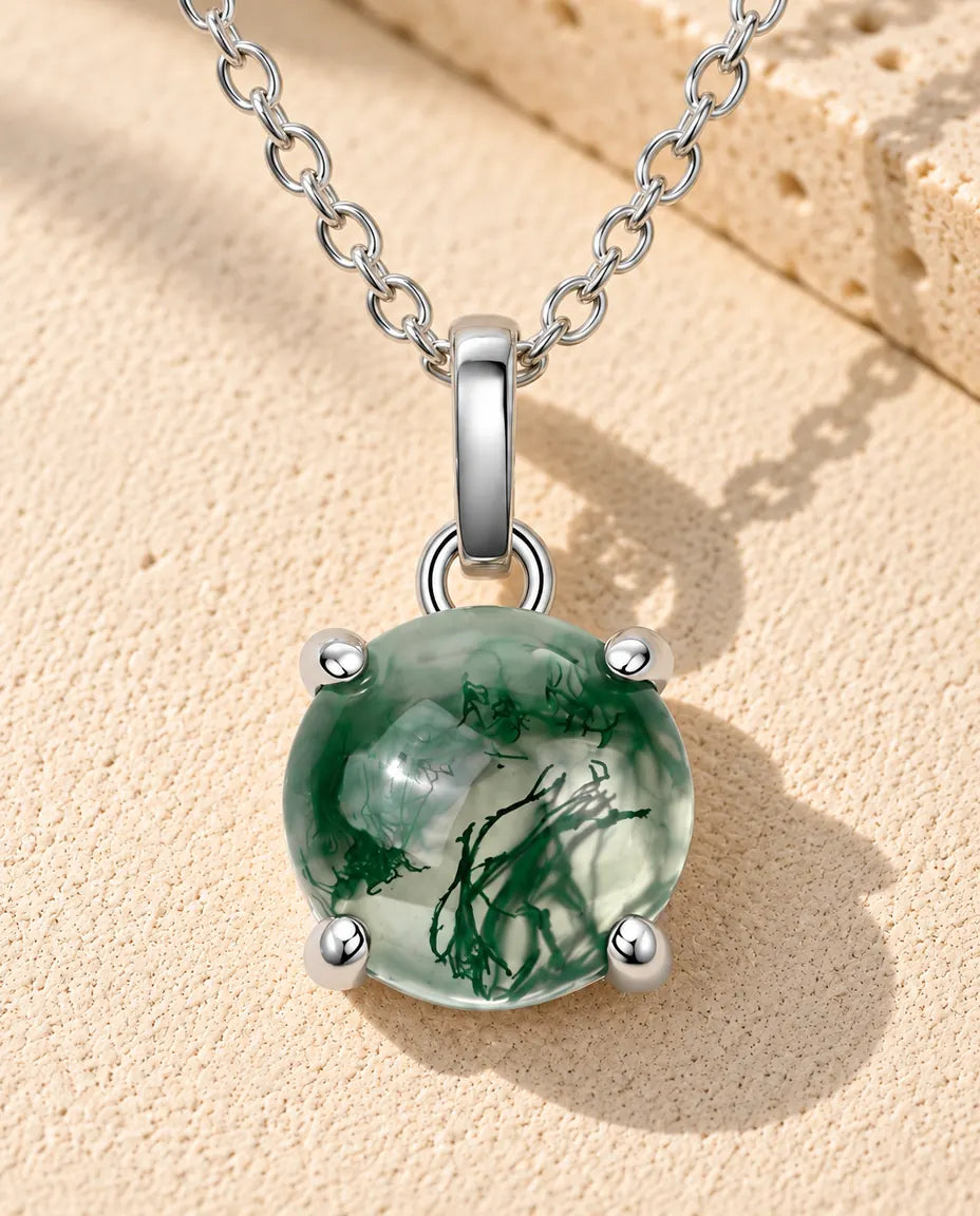 Sølvhalskæde med fire kløer, der holder en rund og højt hvælvet grøn moss agate, på beige puds foran travertin.