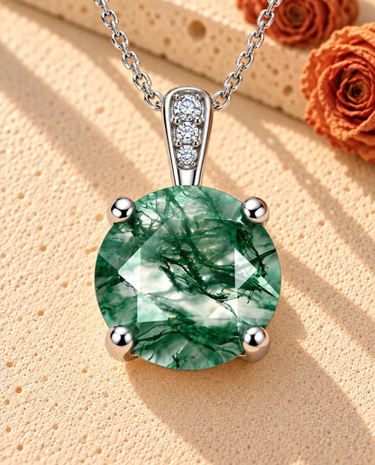 Sølvhalskæde med en rund moss agate i fire kløer og tre små klare sten i øskenen på beige puds foran travertin.