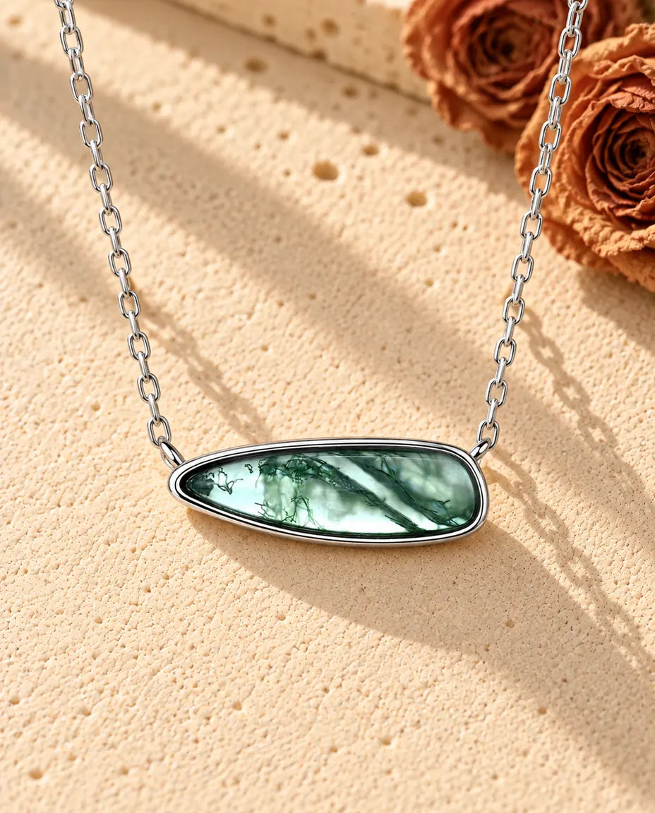 Sølvhalskæde med en lang, glat moss agate-dråbe lagt vandret i en enkel fatning på beige puds foran travertin.