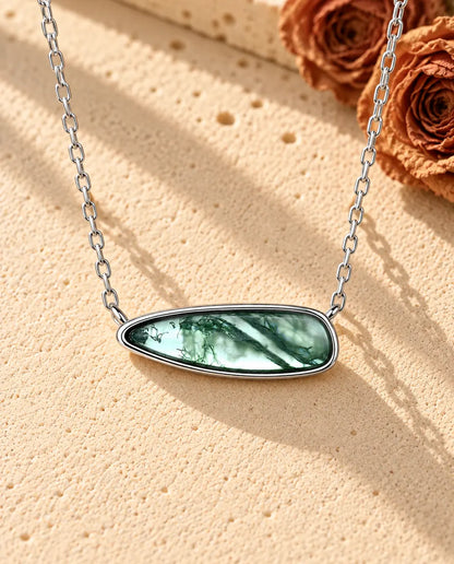 Sølvhalskæde med en lang, glat moss agate-dråbe lagt vandret i en enkel fatning på beige puds foran travertin.