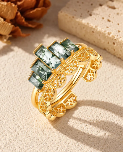 To stablede guldbelagte ringe, den ene med fem baguetteslebne moss agate-sten, på beige puds foran travertin og en rose.
