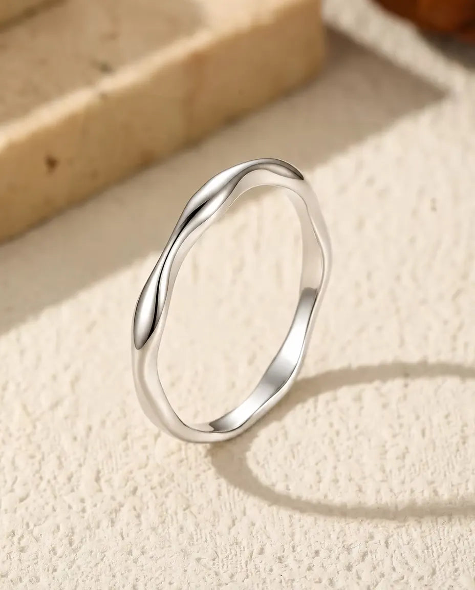 Smal bølget ring i poleret 925 sterling sølv, stillet skråt op på råhvid puds med en buet skygge foran lyse travertintrin.