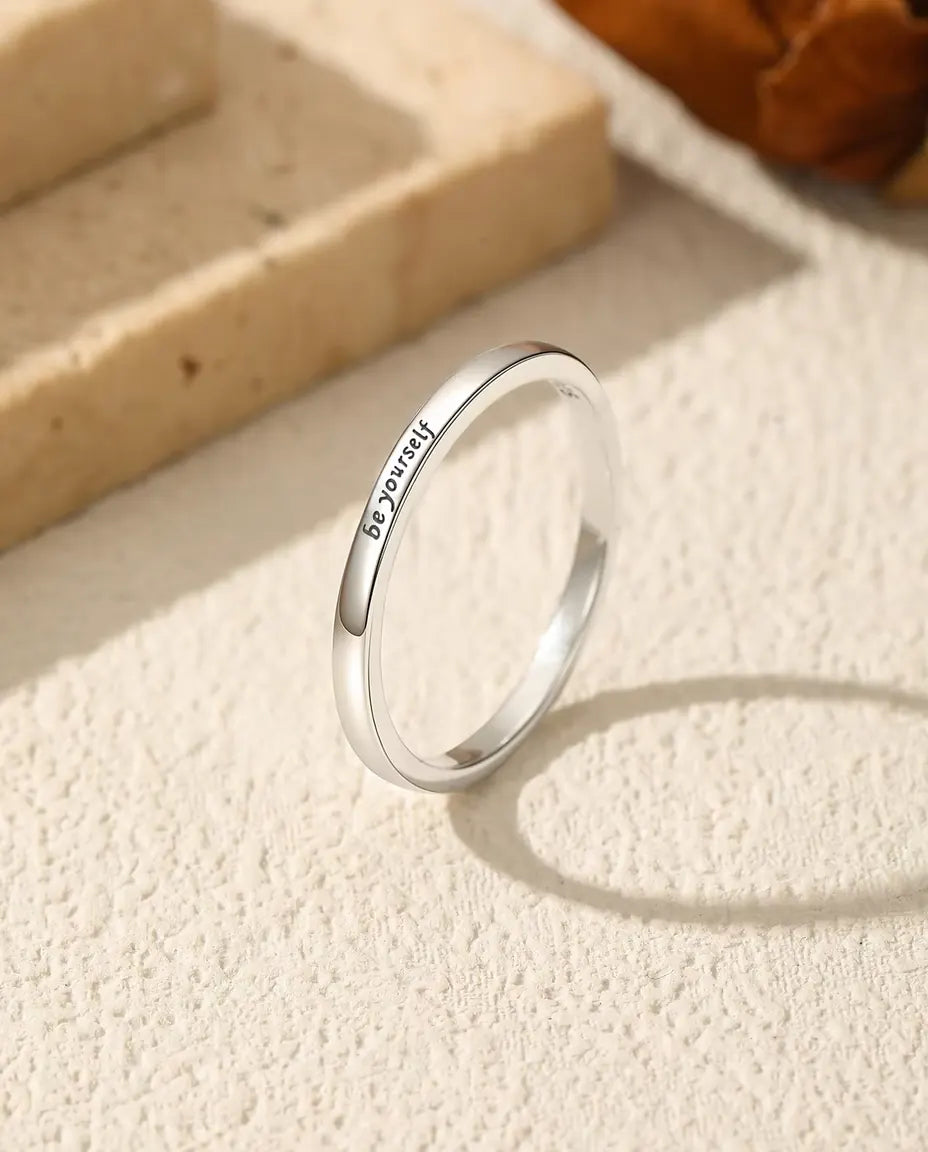 Tynd poleret ring i 925 sterling sølv med de små ord be yourself står på groft puds med oval skygge ved travertin og blomst.