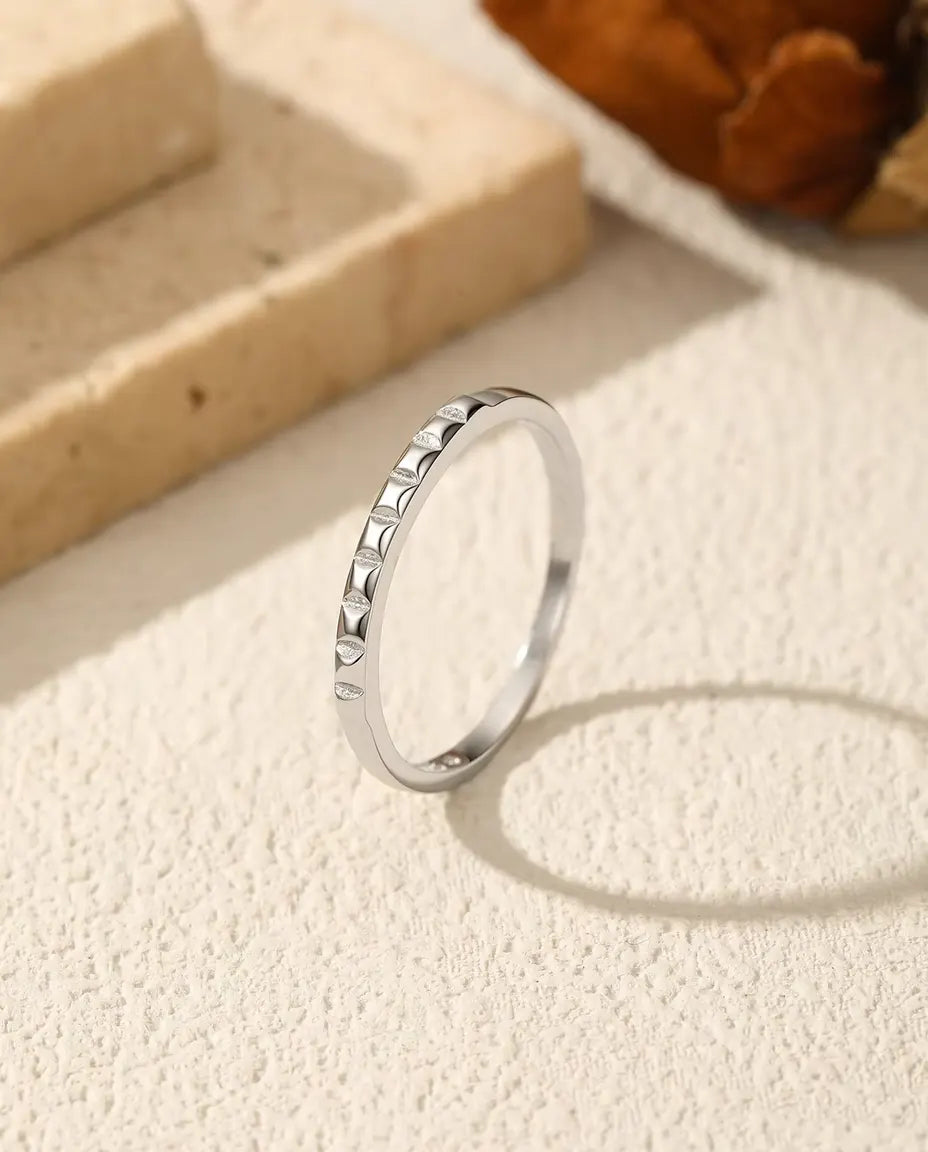 Tynd ring i 925 sterling sølv med små pyramidenitter foran står på groft puds med oval skygge ved travertin og tørret blomst.